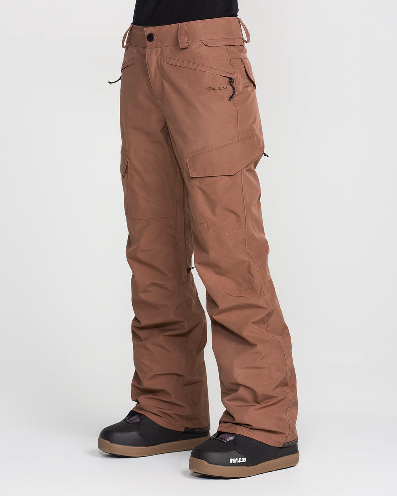 A woman wearing Volcom Women’s Stonelark Gore-Tex Pants in brown stands against a plain white background. The waterproof pants feature side pockets and a slightly loose fit, making them ideal for winter or outdoor activities. She pairs them with black snow boots.