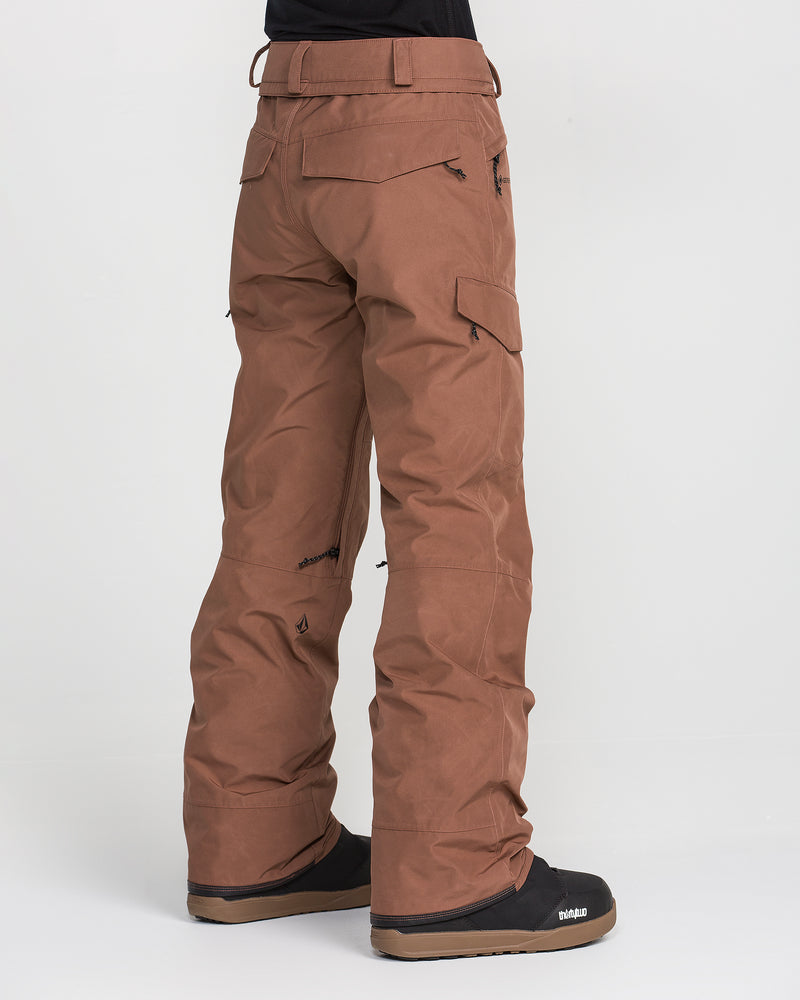 A person wearing Volcom Women’s Stonelark Gore-Tex Pants in a brown cargo style and black snow boots stands against a plain white background. The image is taken from behind, highlighting the pocket and seam details on the women’s waterproof pants.