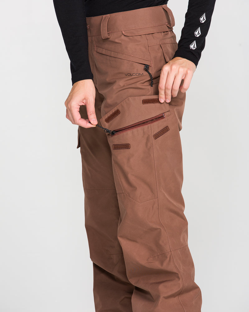 A person is wearing Volcom Women’s Stonelark Gore-Tex Pants in brown, which feature multiple zippered pockets and distinctive black VOLCOM lettering. One hand is pulling a thigh zipper, highlighting the pants’ functional design. Paired with a black long sleeve top with logo accents, this look combines durability, style, and winter-ready performance.