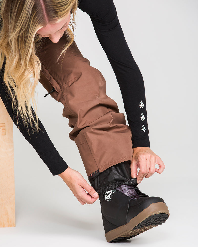 A person wearing Volcom Women’s Stonelark Gore-Tex Pants in brown and a black long-sleeve shirt adjusts the cuff of their pants over a black snow boot.