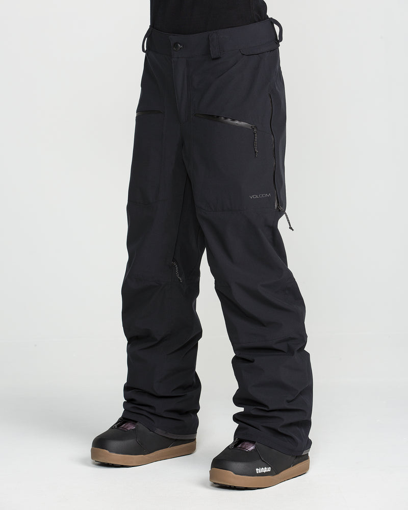 A person is shown from the waist down, wearing Volcom Women’s Karat Stretch 30K Pants in black—featuring 4-way stretch fabric and zippered pockets—paired with black snow boots, standing against a plain white background.
