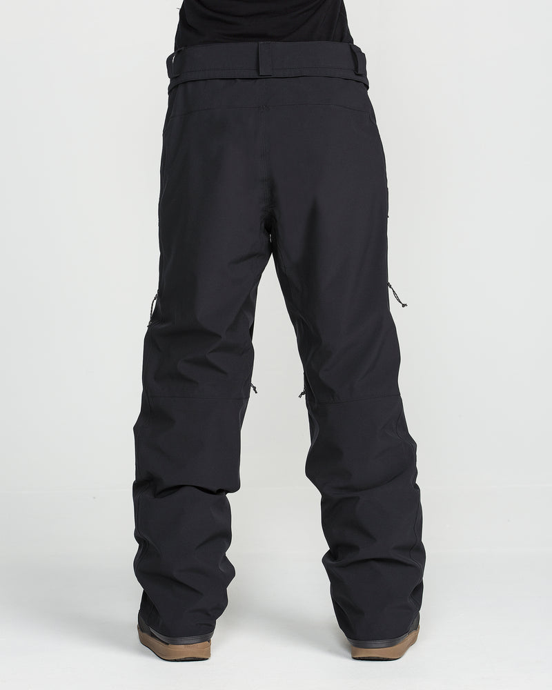 A person is shown from the waist down wearing Volcom Women’s Karat Stretch 30K Pants in black—loose-fitting, waterproof pants made from recycled fabric—paired with brown boots, standing against a plain white background.