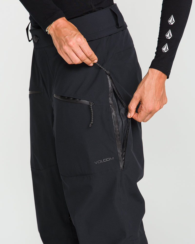 A person unzips a pocket on black Volcom Women’s Karat Stretch 30K Pants, revealing the design and waterproof zipper details enhanced by V-Science 2-Layer construction. Only their torso and arms are visible, and they are wearing a black long-sleeve shirt.