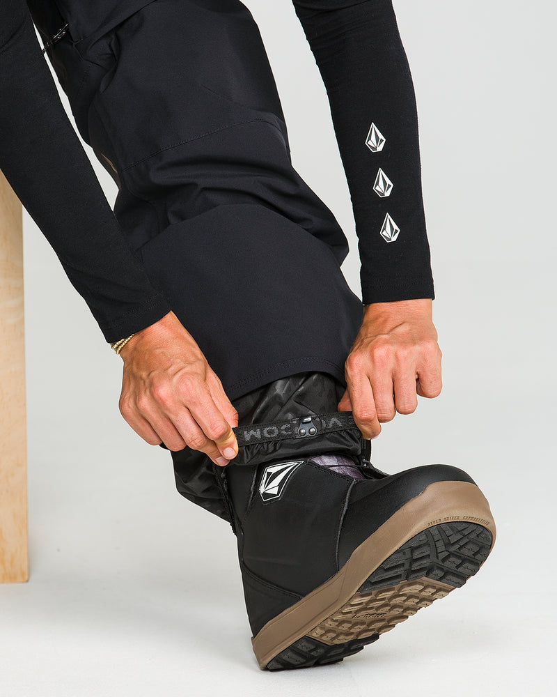 A close-up shows a person adjusting the leg of their Volcom Women’s Karat Stretch 30K Pants over black boots, with the Volcom logo visible on the pants.