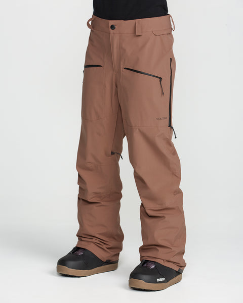 A person is wearing Volcom Women’s Karat Stretch 30K Pants in brown, featuring black zippers and paired with black boots, standing against a plain white background. Only the lower half of their body is visible.