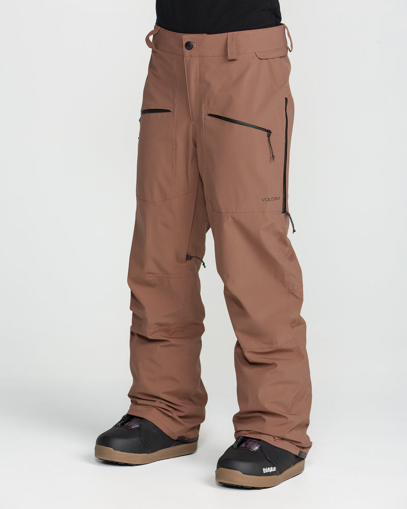 A person is wearing Volcom Women’s Karat Stretch 30K Pants in brown, featuring black zippers and paired with black boots, standing against a plain white background. Only the lower half of their body is visible.