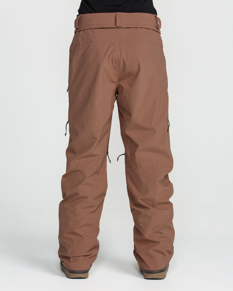 A person wearing the Volcom Women’s Karat Stretch 30K Pants in brown, made from recycled polyester ski pant material, paired with brown boots, is standing against a plain white background. The pants feature zippered side pockets and offer a loose, relaxed fit. The upper body is not visible.