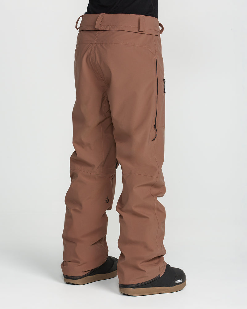 A person is wearing Volcom Women’s Karat Stretch 30K Pants in brown, paired with black boots, standing against a white background. The relaxed-fit ski pants feature visible seams, belt loops, and a side zipper pocket. Only the lower half of the body is shown.