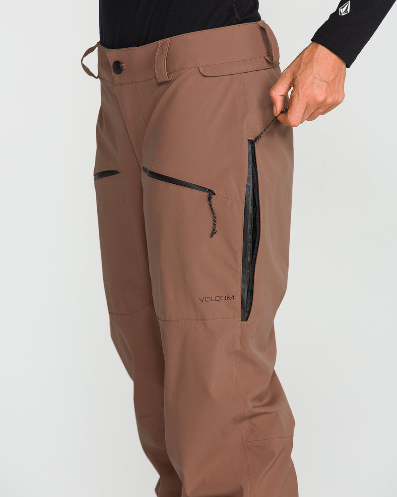 A person dressed in a black long-sleeve shirt wears Volcom Women’s Karat Stretch 30K Pants, crafted from recycled polyester ski pant material. The brown waterproof ski pants feature black zippers, a button closure, and the person is seen opening one of the zippered side pockets.