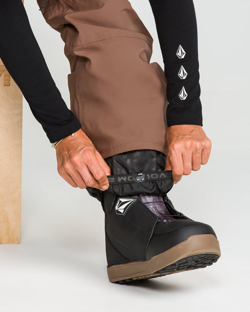 Wearing Volcom Women’s Karat Stretch 30K Pants in brown and a black long-sleeve top, a person sits on a wooden bench adjusting the cuff of their pants over a black snow boot with a visible logo. Their hands are clearly shown.