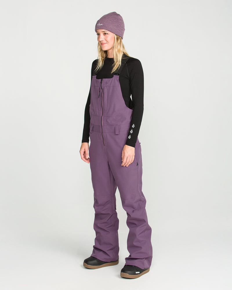 A woman wearing a purple beanie, black long-sleeve shirt, and Volcom Women’s Swift Bib waterproof snow pants made from recycled fabric stands facing slightly left in a studio with a plain white background.