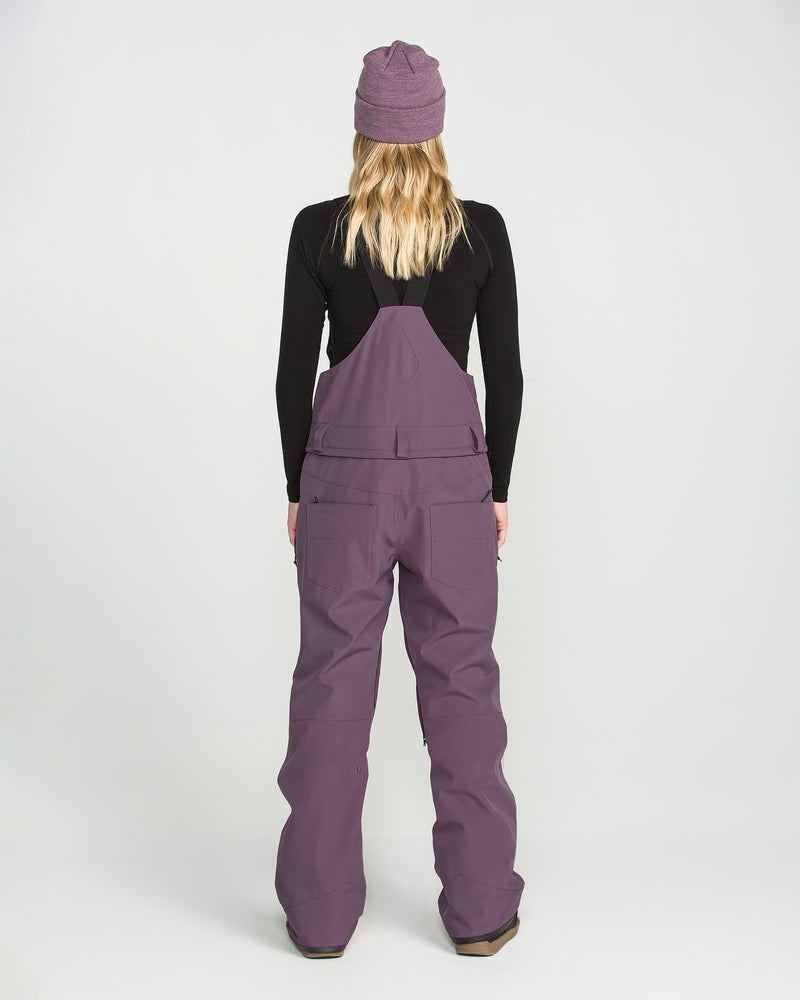 A person with long blonde hair, wearing a purple beanie, a black long-sleeve top, and the Volcom Women’s Swift Bib in purple—crafted from recycled fabric—stands facing away from the camera against a plain white background.