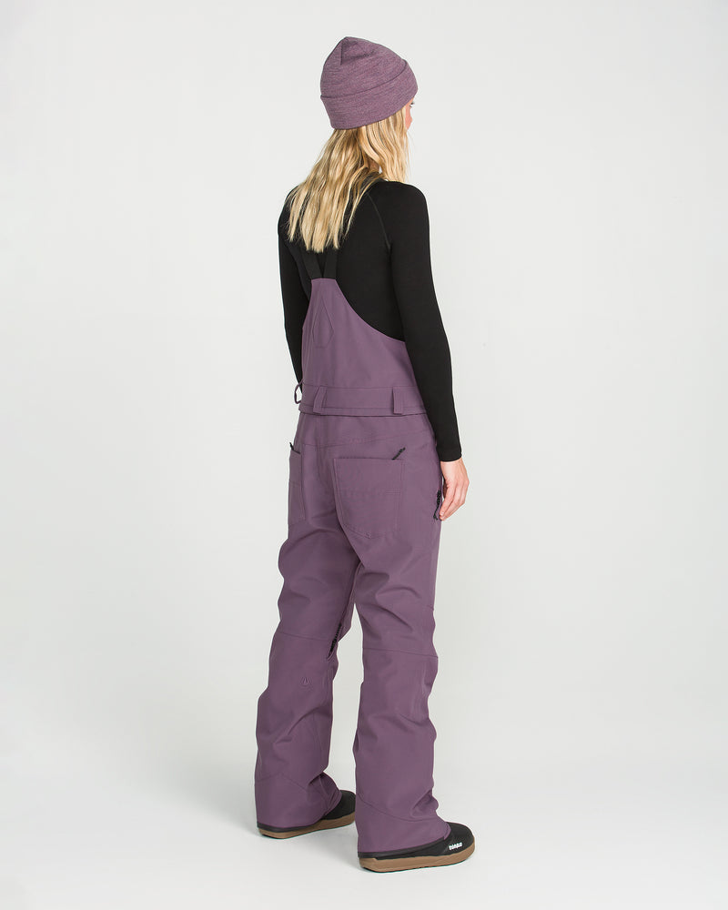 A person with long blonde hair, wearing a purple beanie, black top, and the Volcom Women’s Swift Bib in purple recycled fabric, stands facing away from the camera against a plain white background.