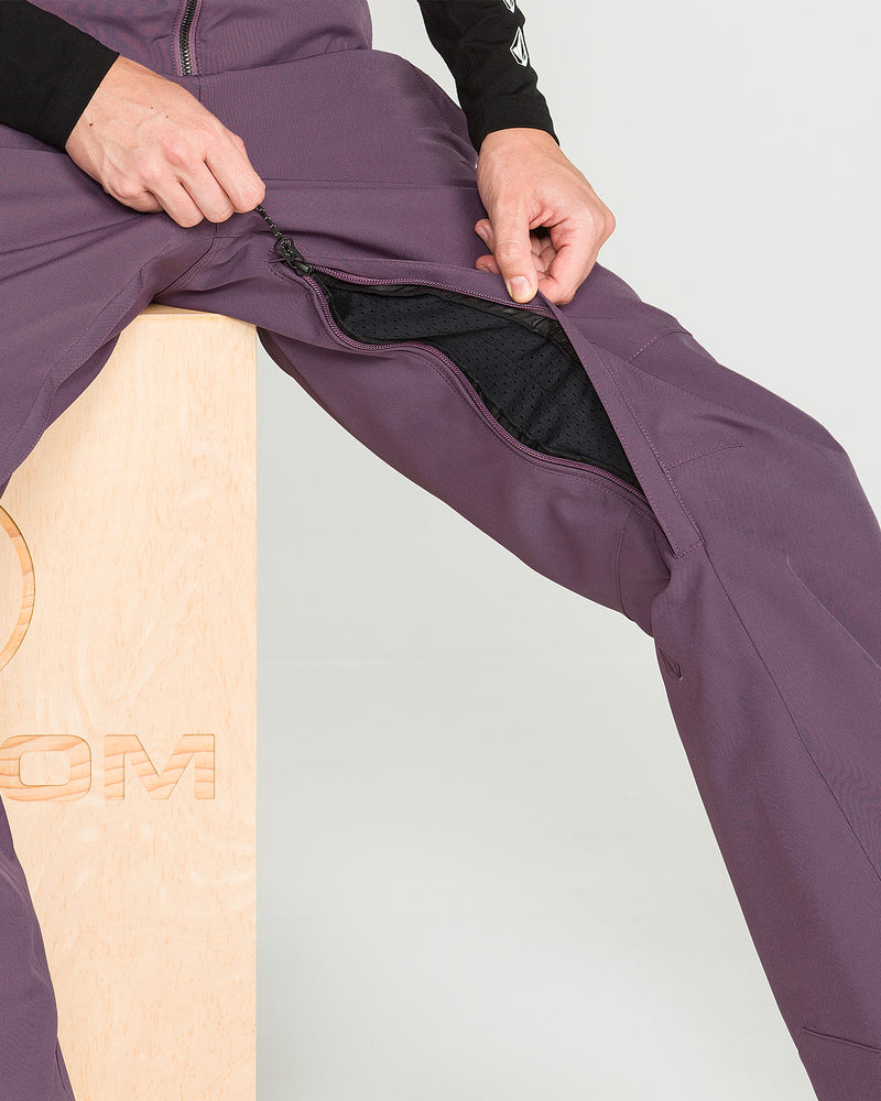 A person sits on a wooden box wearing Volcom Women’s Swift Bib purple waterproof snow pants and unzips a large zipper along the inner thigh, revealing a black mesh lining inside.