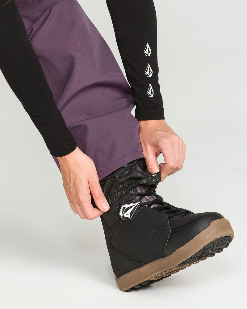 A person fastens the laces of a black snowboarding boot while wearing the Volcom Women’s Swift Bib in a vibrant purple and a black long-sleeve shirt featuring white sleeve logos. The background is plain and light-colored.