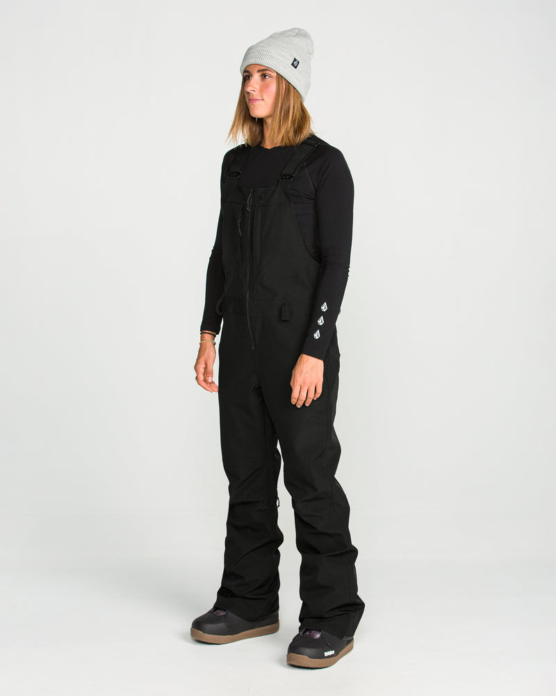 A person stands facing slightly left against a plain white background, wearing a gray beanie, black long-sleeve shirt, and the Volcom Women’s Swift Bib—sleek black women’s overalls made from V-Science stretch recycled fabric.