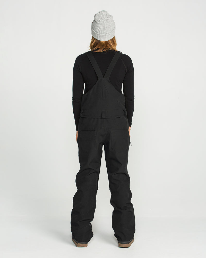 A person stands facing away from the camera against a plain white background, wearing a gray beanie, black long-sleeve shirt, and the Volcom Women’s Swift Bib made from recycled fabric.