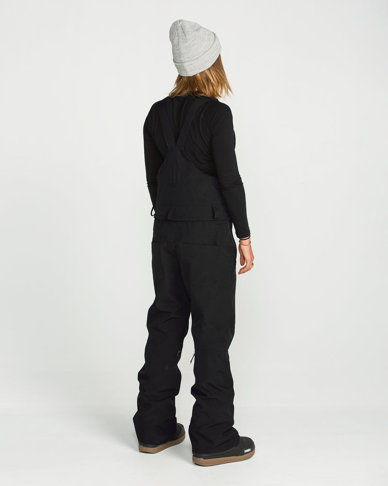 A person stands facing away, wearing a gray beanie, a black long-sleeve shirt, and the Volcom Women’s Swift Bib made from V-Science stretch recycled fabric, paired with black pants and brown-soled shoes against a plain white background.