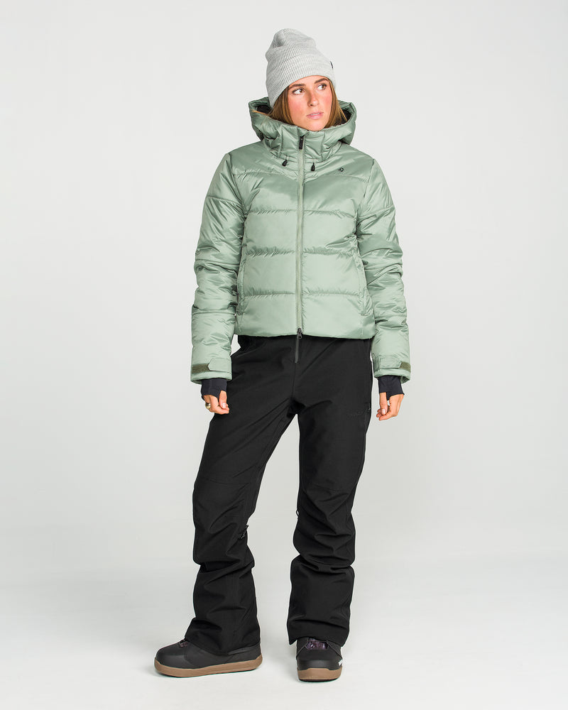 A person wears the Volcom Women’s Swift Bib paired with a light green recycled-fabric puffer jacket, gray beanie, and winter boots, standing against a plain white background.