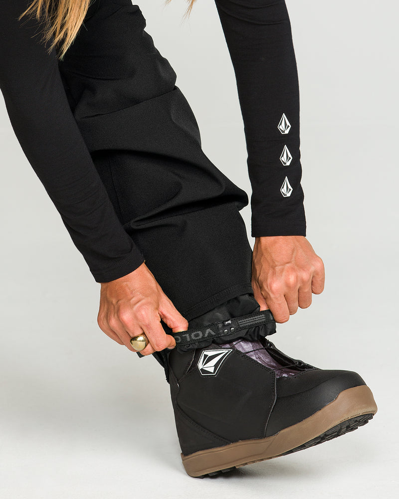 A person wearing Volcom’s Women’s Swift Bib in black—crafted from recycled fabric—and a black long-sleeve shirt, each featuring a white geometric Volcom logo, adjusts the cuff over a black snow boot. The person’s hands are visible, with one hand adorned by a gold ring.
