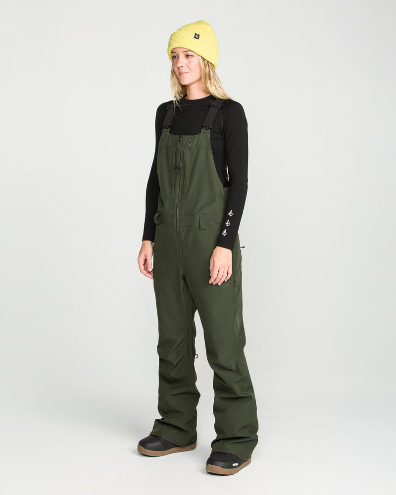 A woman wearing a yellow beanie, black long-sleeve shirt, and the Volcom Women’s Swift Bib—crafted from recycled fabric—stands against a plain white background, looking slightly to the side.