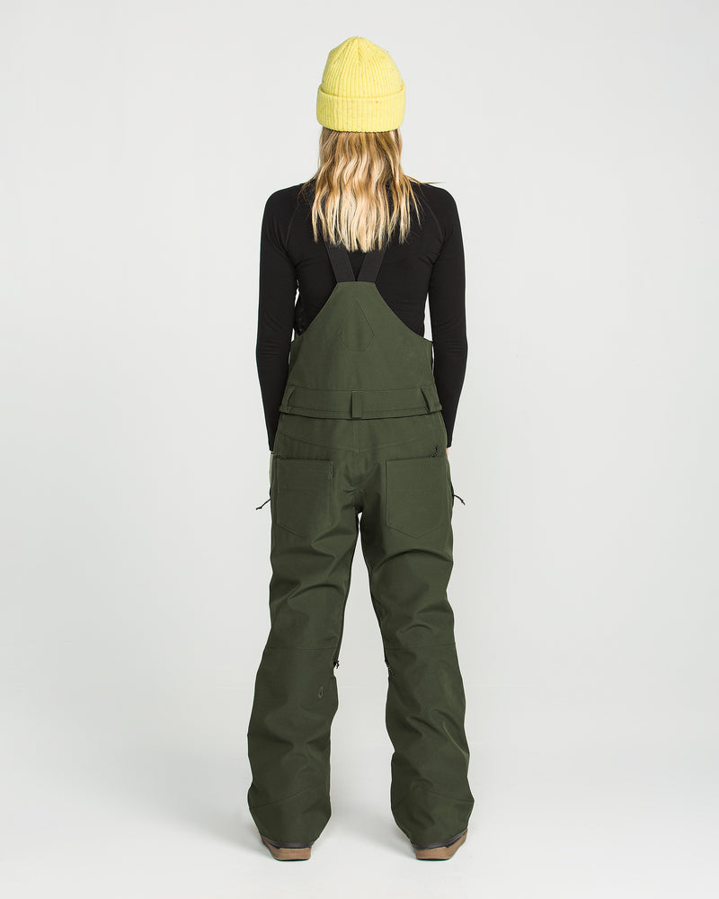 A person stands facing away, wearing a yellow beanie, a black long-sleeve shirt, and the Volcom Women’s Swift Bib—green wide-leg overalls with large back pockets made from recycled fabric. The background is plain white.