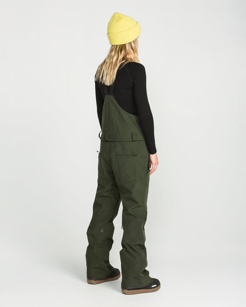 A person with long blonde hair is seen from behind against a plain white background, wearing a yellow beanie, a black long-sleeve top, and the Volcom Women’s Swift Bib in green, made from recycled fabric.