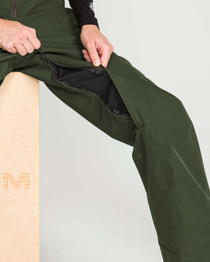 A person sitting on a wooden box unzips the side of their Volcom Women’s Swift Bib in dark green, revealing an inner black layer. Only their lower torso, hands, and legs are visible.