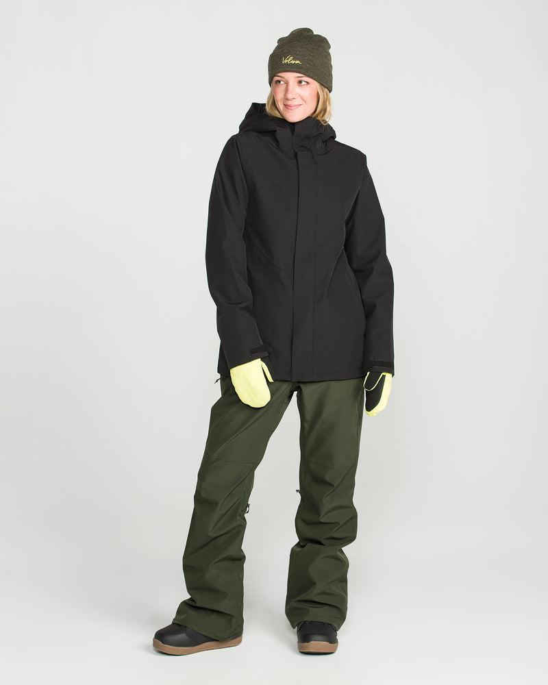 A woman wearing the Volcom Women’s Swift Bib stands on a white background, smiling slightly and looking to the side. She pairs the bib with a black winter jacket, yellow gloves, black boots, and a green beanie.