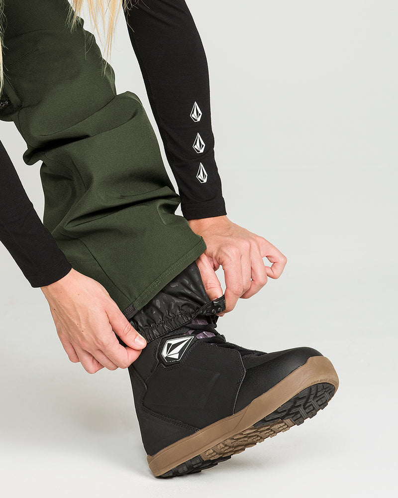 A person wearing the Volcom Women’s Swift Bib—a pair of green waterproof snow pants featuring white geometric logo designs—tightens the laces on a black snowboarding boot with a brown sole while dressed in a long-sleeved shirt.