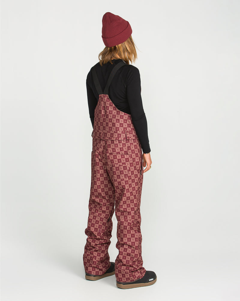A person stands facing away, wearing a maroon beanie, black long-sleeve shirt, and the Volcom Women’s Swift Bib—maroon patterned overalls crafted from recycled fabric—with black boots, set against a plain white background.