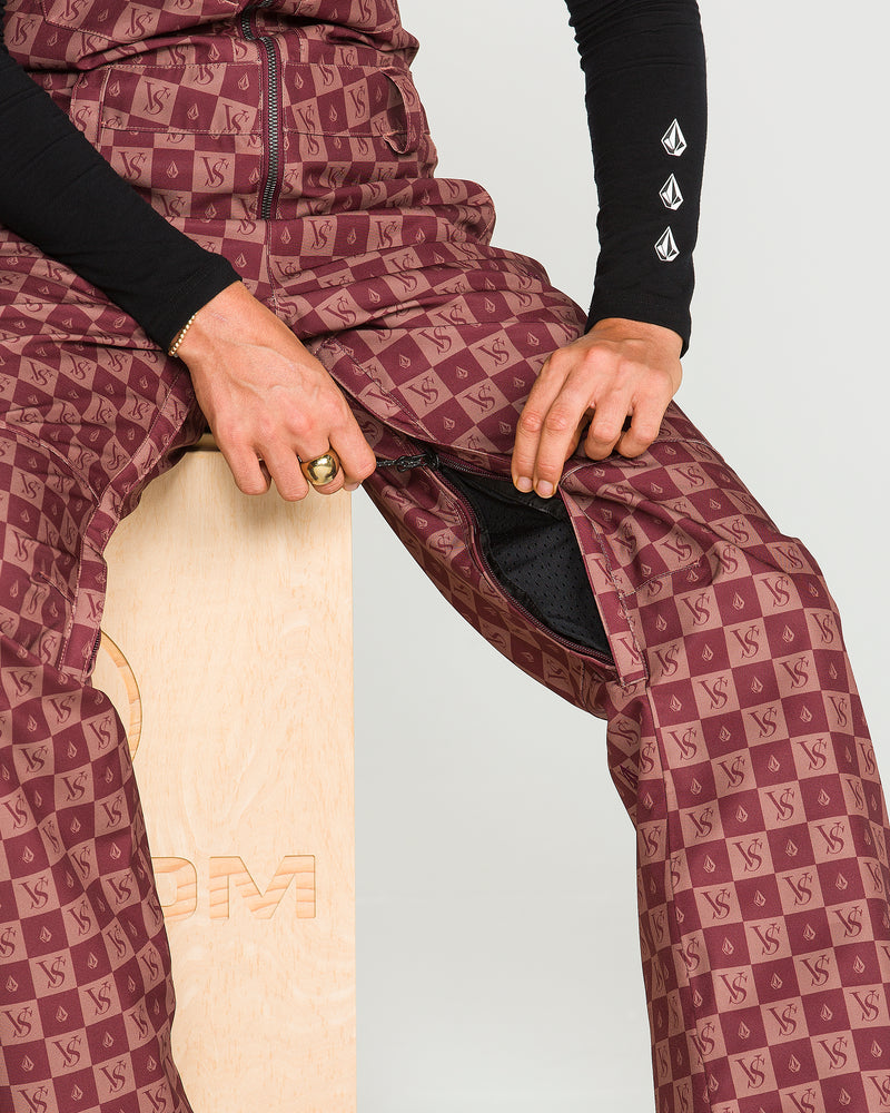 A person sits on a wooden block unzipping the Volcom Women’s Swift Bib, crafted from recycled fabric in a maroon pattern with a repeating logo, paired with a black long-sleeve shirt featuring white sleeve symbols and accessorized with a gold ring.