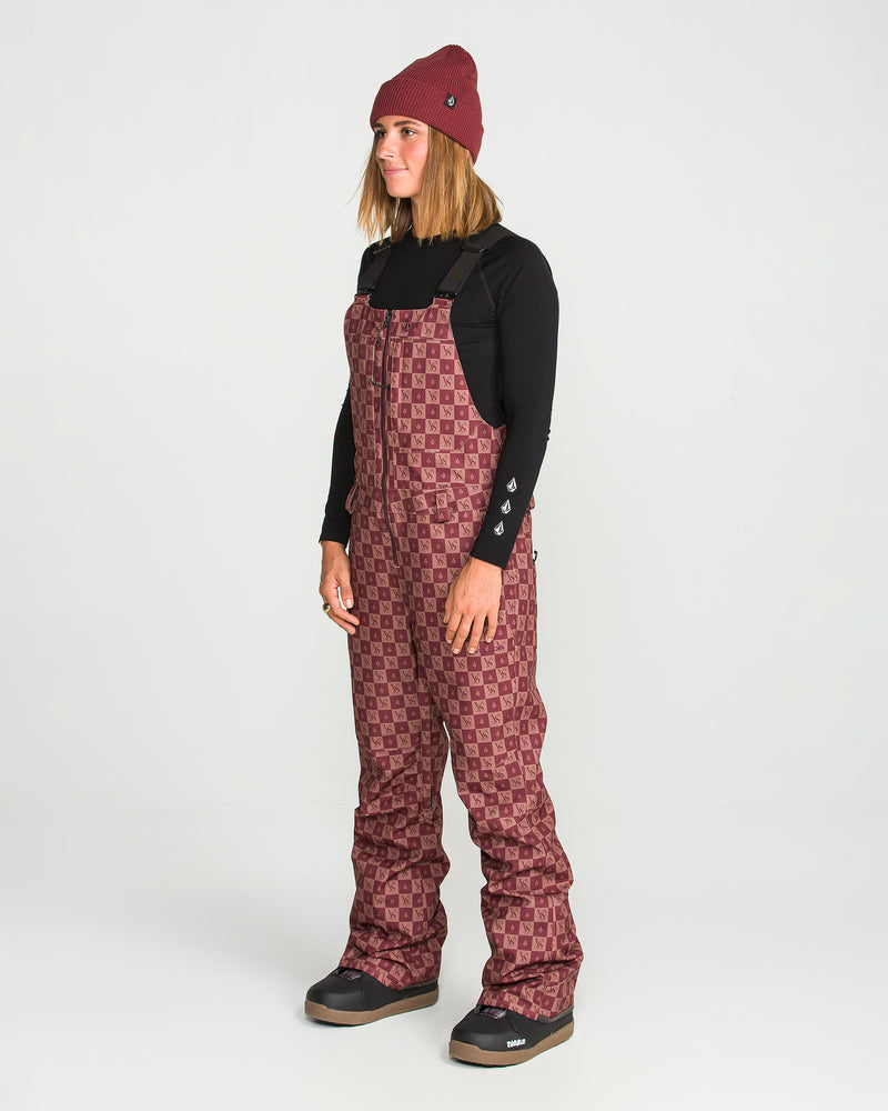 Wearing a maroon beanie, a black long-sleeve shirt, and Volcom’s Women’s Swift Bib in a patterned maroon design made from recycled fabric, a person stands facing slightly left against a plain white background.