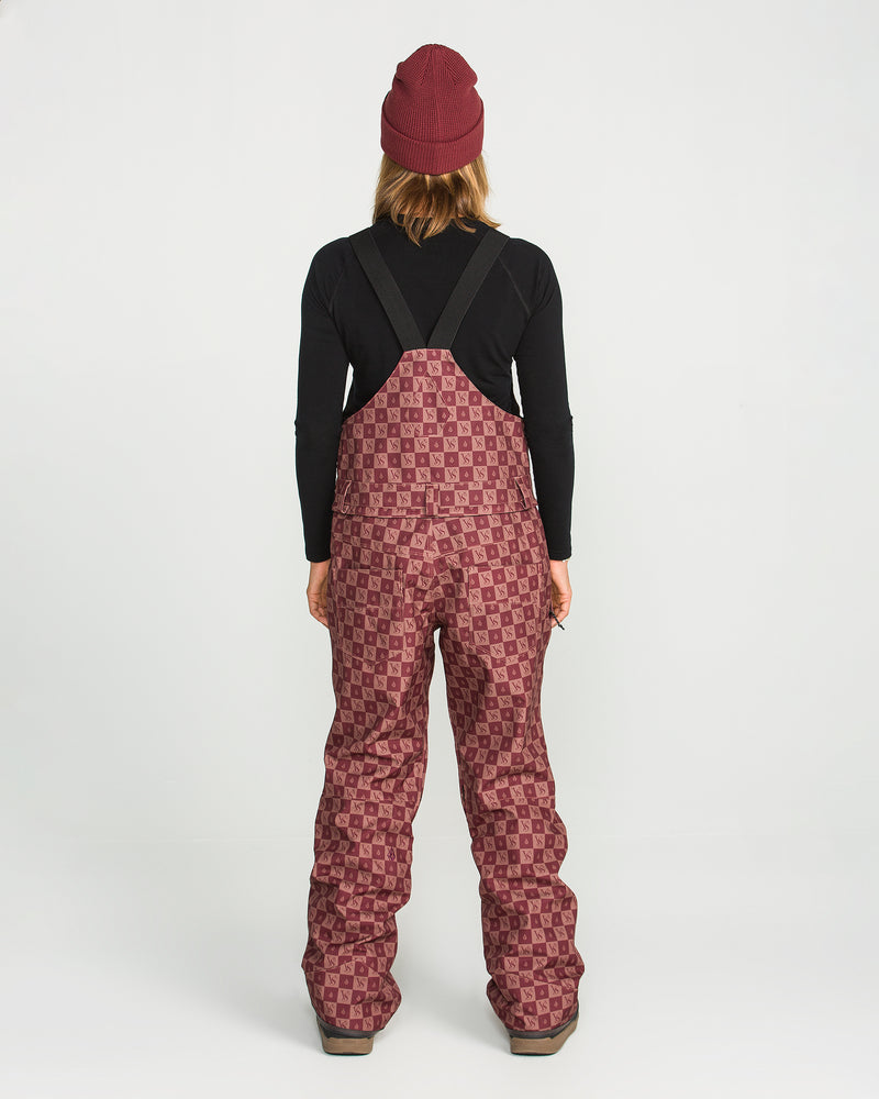 A person facing away, wearing a maroon beanie, a black long-sleeve shirt, and Volcom Women’s Swift Bib overalls in a maroon pattern with black straps—crafted from recycled fabric and featuring V-Science stretch—stands against a plain white background.