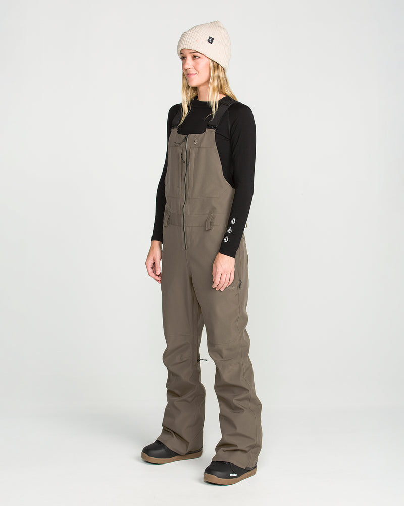 A person stands facing slightly left on a plain white background, wearing a beige beanie, a black long-sleeve top, and the Volcom Women’s Swift Bib in brown, made from recycled fabric.