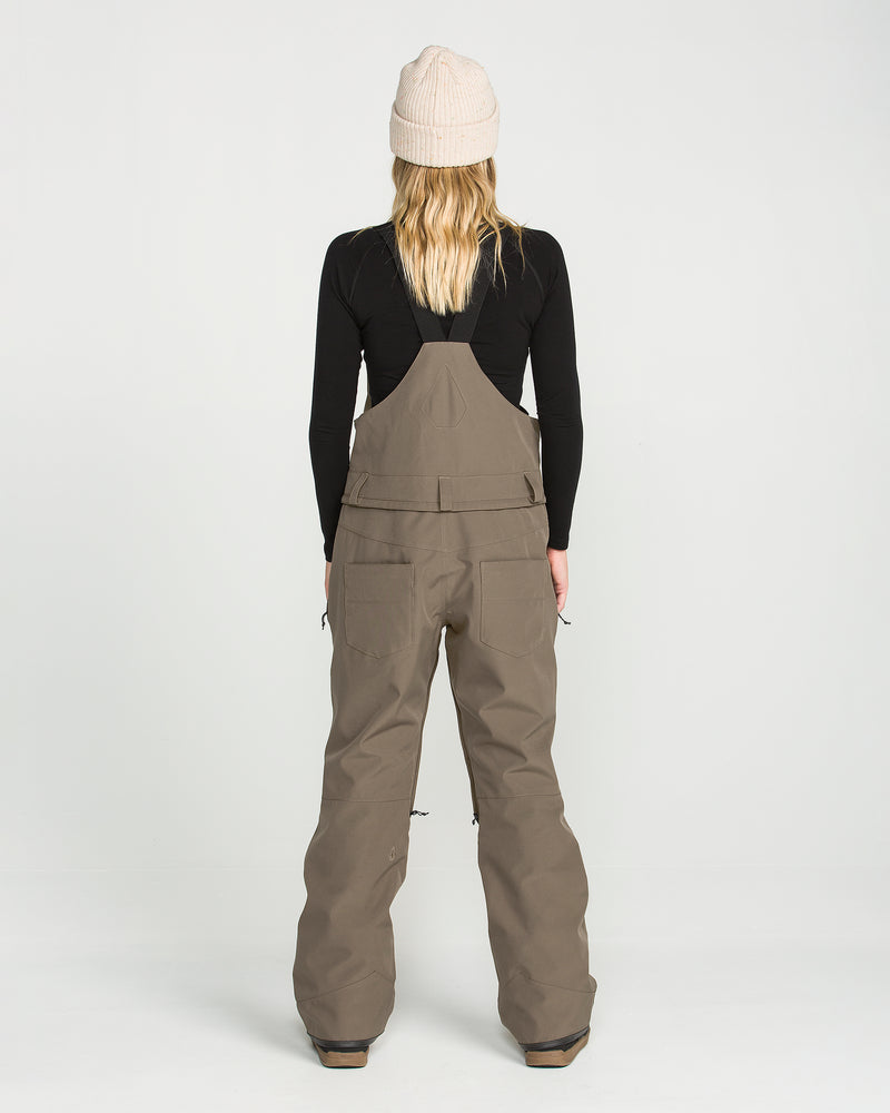 A person wearing the Volcom Women’s Swift Bib in tan—crafted from recycled fabric—paired with a black long-sleeve shirt and a beige knit beanie, stands facing away from the camera against a plain white background.