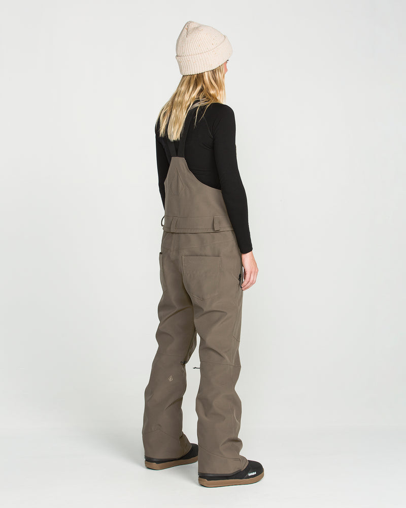 A person with long blonde hair, wearing a beige beanie, black long-sleeve top, and the Volcom Women’s Swift Bib in taupe—crafted from recycled fabric—stands facing away on a plain white background.