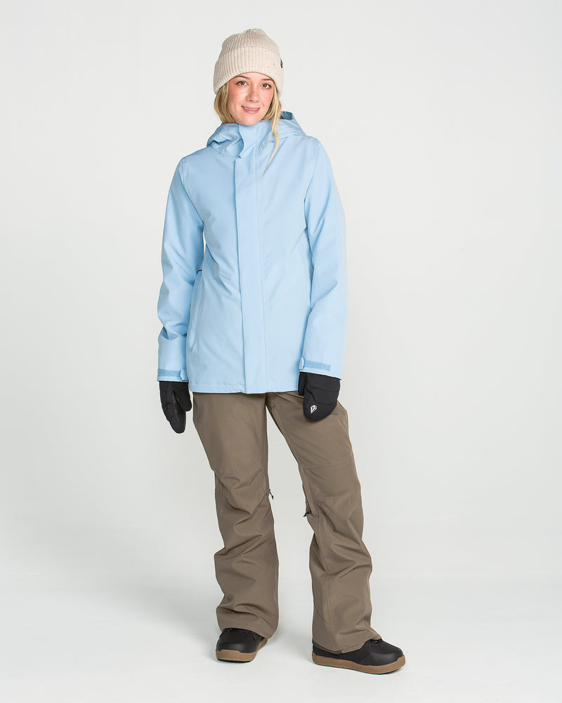 A person stands against a plain white background, smiling at the camera and wearing the Women’s Swift Bib by Volcom—a light blue jacket made from recycled fabric, paired with a beige beanie, black gloves, brown pants, and black boots.