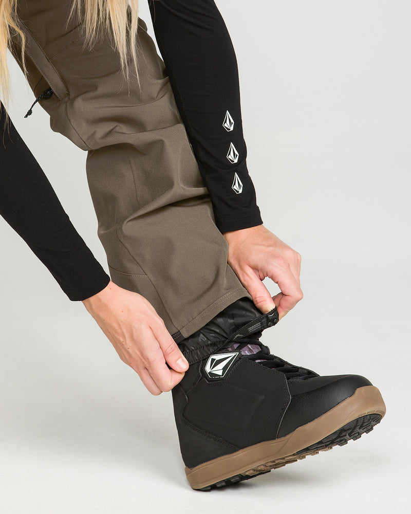 A person tucks the brown Volcom Women’s Swift Bib waterproof overalls into a black snowboarding boot. They wear a black long-sleeve shirt with white geometric logos on the sleeve, standing against a plain, light-colored background.