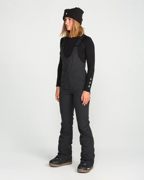 A woman wearing a black beanie, long-sleeve shirt, and Volcom Women’s Battle Stretch Bib—featuring PFAS-free DWR, 15K/10K protection, and stretch fabric—stands on a white background looking slightly to the side with a relaxed expression.