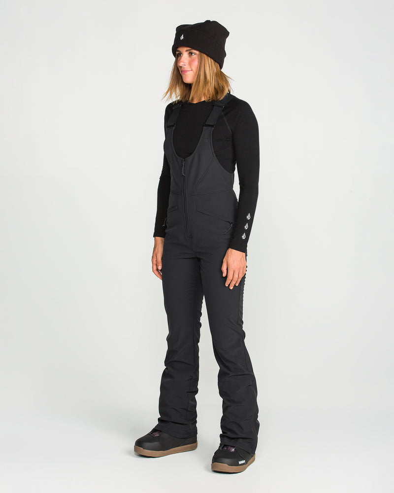 A woman wearing a black beanie, long-sleeve shirt, and Volcom Women’s Battle Stretch Bib—featuring PFAS-free DWR, 15K/10K protection, and stretch fabric—stands on a white background looking slightly to the side with a relaxed expression.