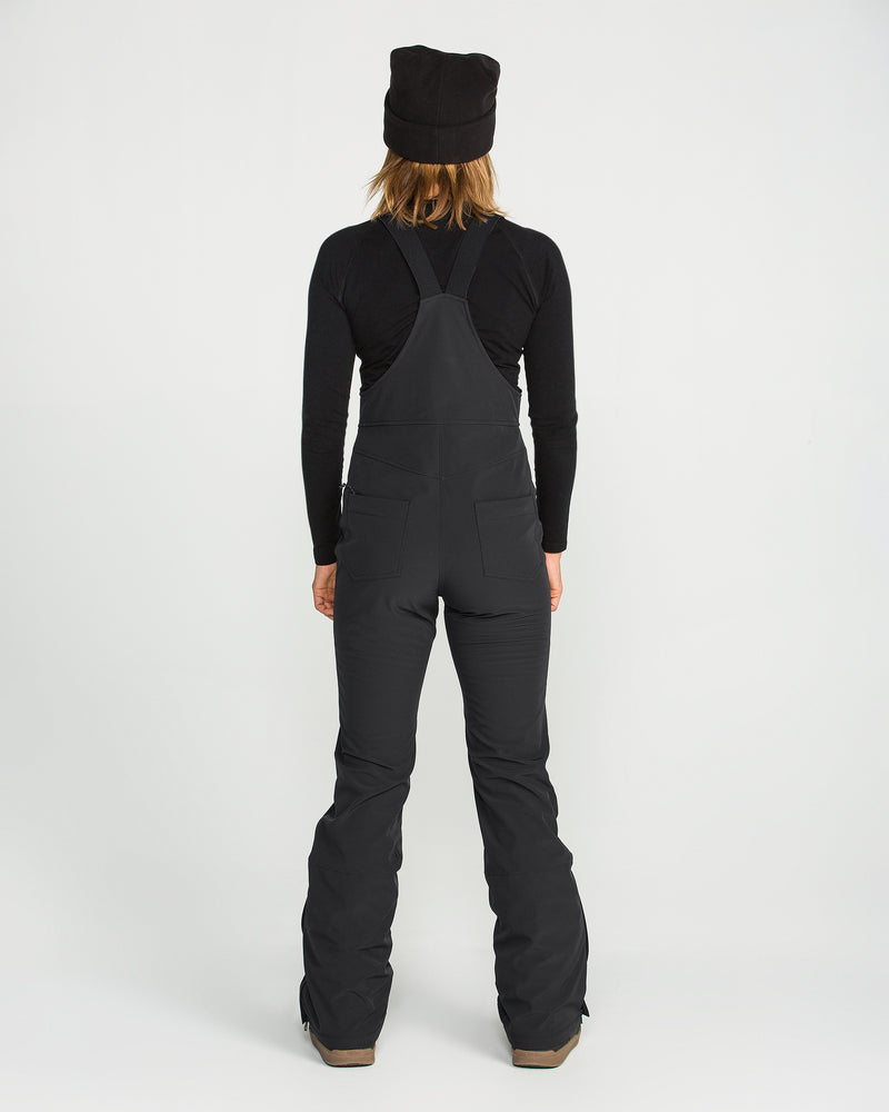 A person facing away, dressed in a black long-sleeve shirt and the Volcom Women’s Battle Stretch Bib—featuring a stretch bib design with PFAS-free DWR for added protection—paired with a black beanie, stands against a plain white background.