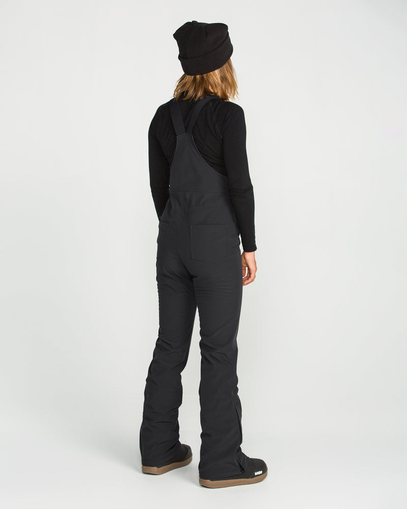 A person stands facing away from the camera against a plain white background, dressed in a black beanie, black long-sleeve shirt, and the Volcom Women’s Battle Stretch Bib in PFAS-free DWR black.