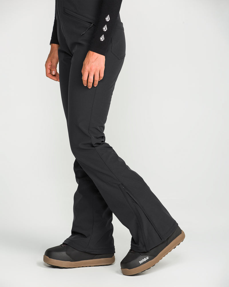 A person is wearing the Volcom Women’s Battle Stretch Bib—black, fitted pants with a slight boot cut at the ankle and PFAS-free DWR for protection against the elements—paired with black winter boots, standing on a white background. Only the lower half of their body is visible.