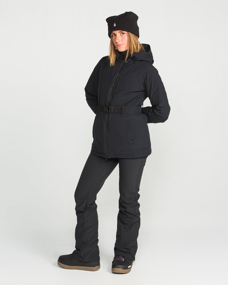 A person stands against a plain background wearing the Volcom Women’s Battle Stretch Bib paired with black boots and a black beanie, hands in pockets and striking a relaxed pose. The outfit features 15K/10K protection and PFAS-free DWR for enhanced weather resistance.