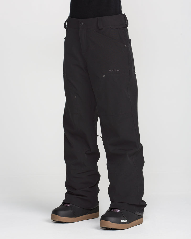 A person wearing Volcom Women’s Pollock Baggy Pants—black, waterproof snow pants made from recycled stretch oxford fabric—paired with black snowboarding boots and standing against a plain white background. These baggy pants feature visible seams, functional pockets, and the Volcom brand logo on the left thigh, combining comfort with sustainable style.