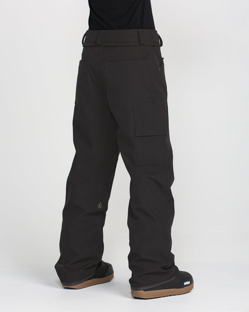 A person stands with their back to the camera against a plain white background, wearing the Volcom Women’s Pollock Baggy Pants—black waterproof snow pants made from recycled stretch oxford material—and black boots with brown soles.