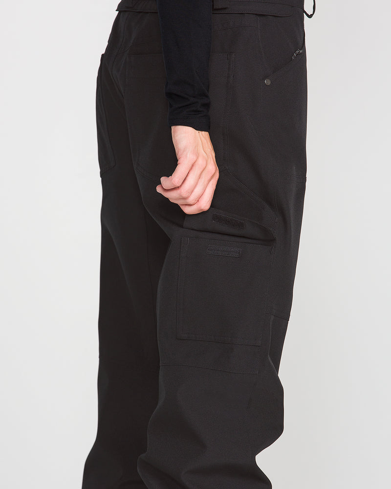 A person is shown from the side wearing the Volcom Women’s Pollock Baggy Pants in black, featuring a loose fit and multiple pockets. Their hand is gripping a seam on the pants. Crafted from recycled stretch oxford fabric, these pants combine comfort with sustainability. The background is plain white.