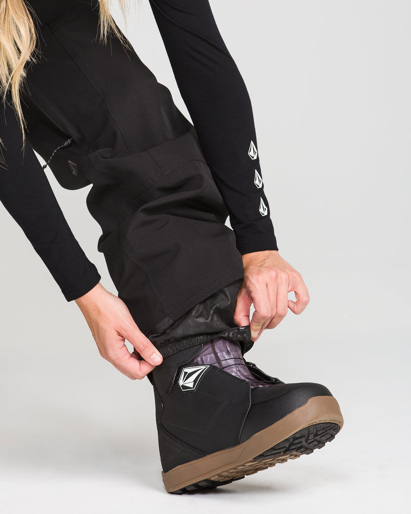 A person adjusts the cuff of Volcom Women’s Pollock Baggy Pants in black recycled stretch oxford over a black snowboarding boot with a brown sole, showing a close-up of hands and lower body against a plain background.