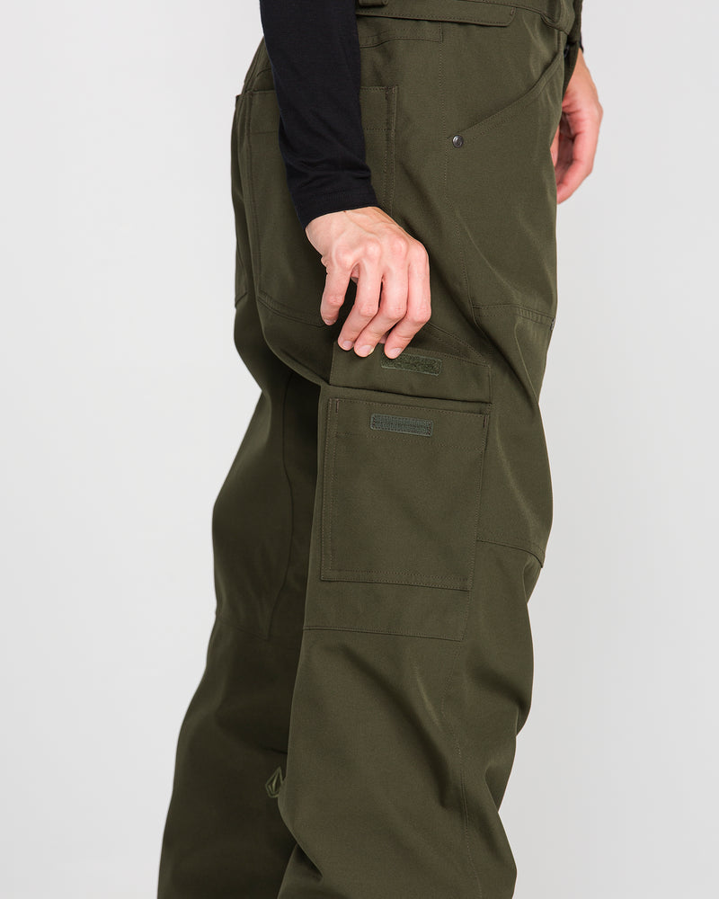 A person wearing Volcom Women’s Pollock Baggy Pants in olive green and a long-sleeve black shirt, shown from the side with one hand resting by their leg.
